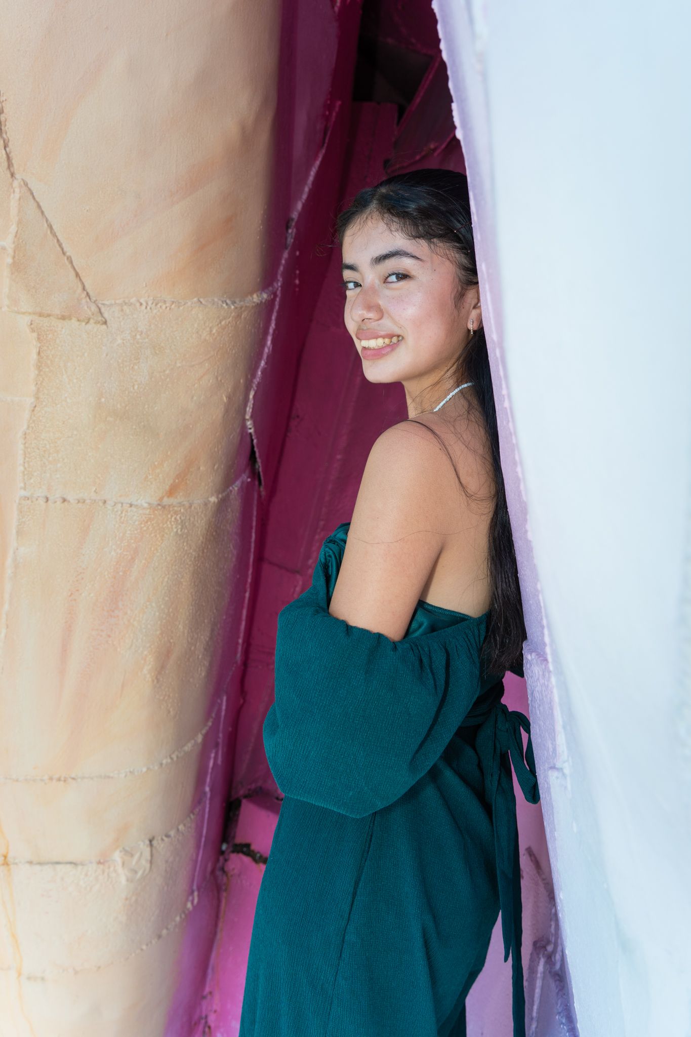 Young woman in teal off-shoulder dress smiling between colorful walls.