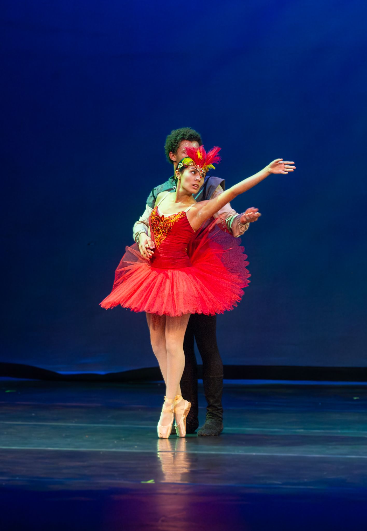 Ballet dancer in red tutu performing graceful pose with partner on dark blue lit stage.