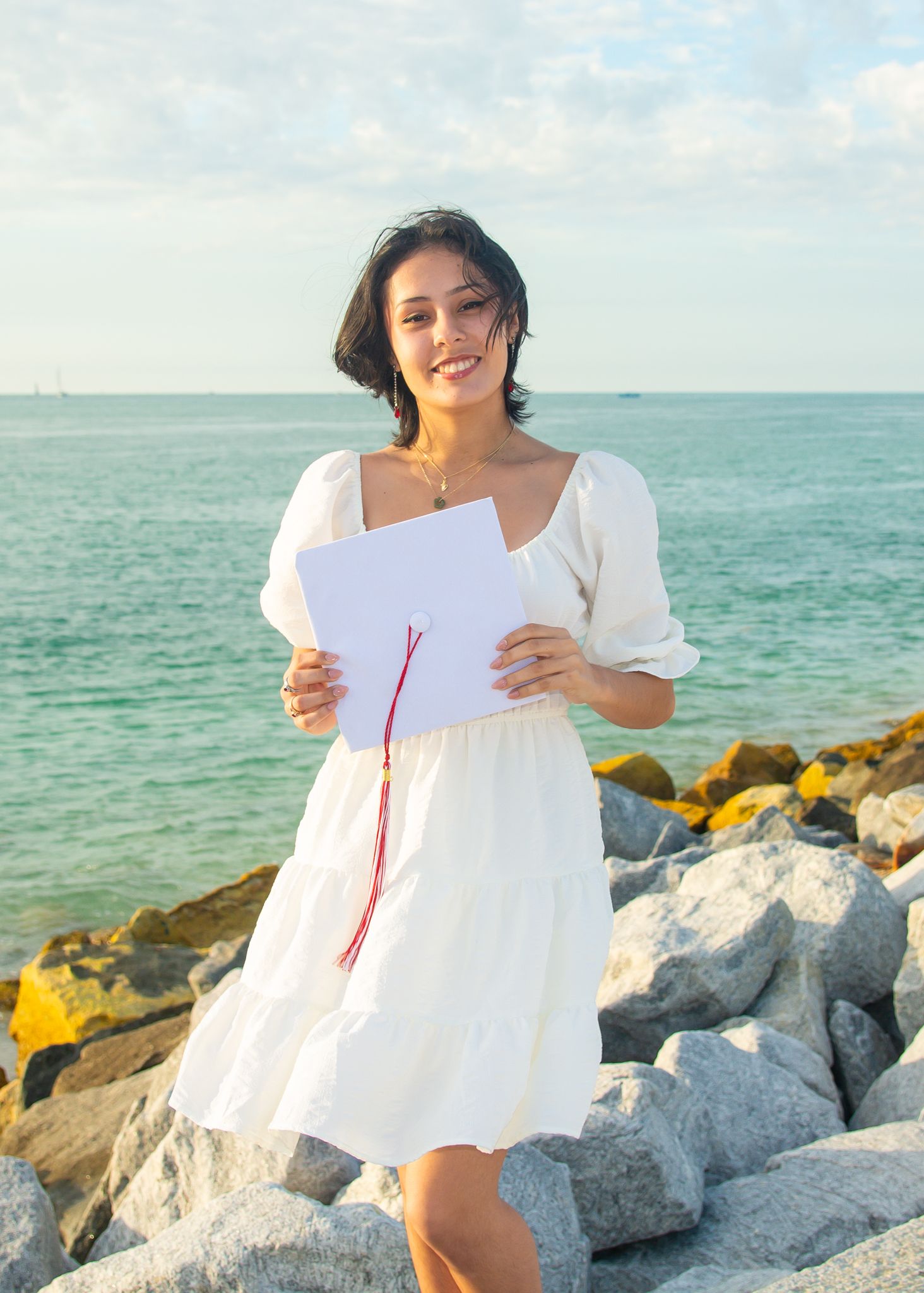 Woman in white sundress holds paper while posing on rocky oceanfront.