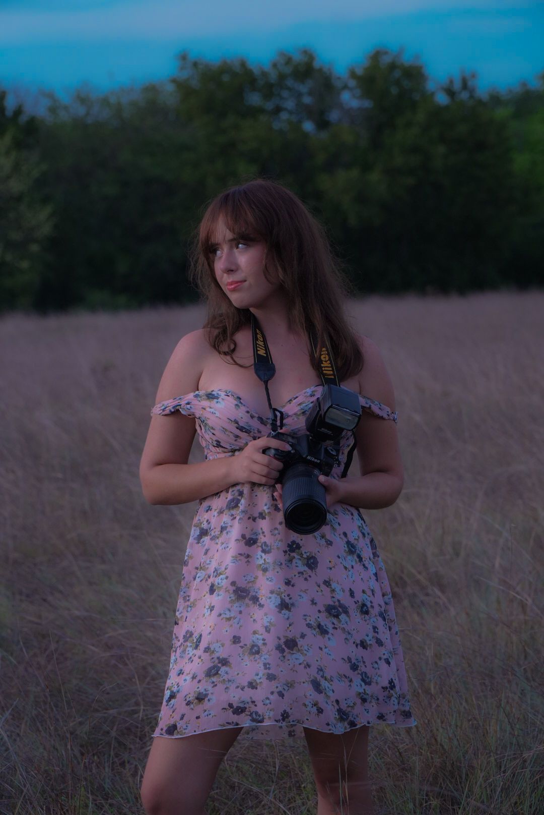 Person in floral dress poses with camera in tall grass field during golden hour.