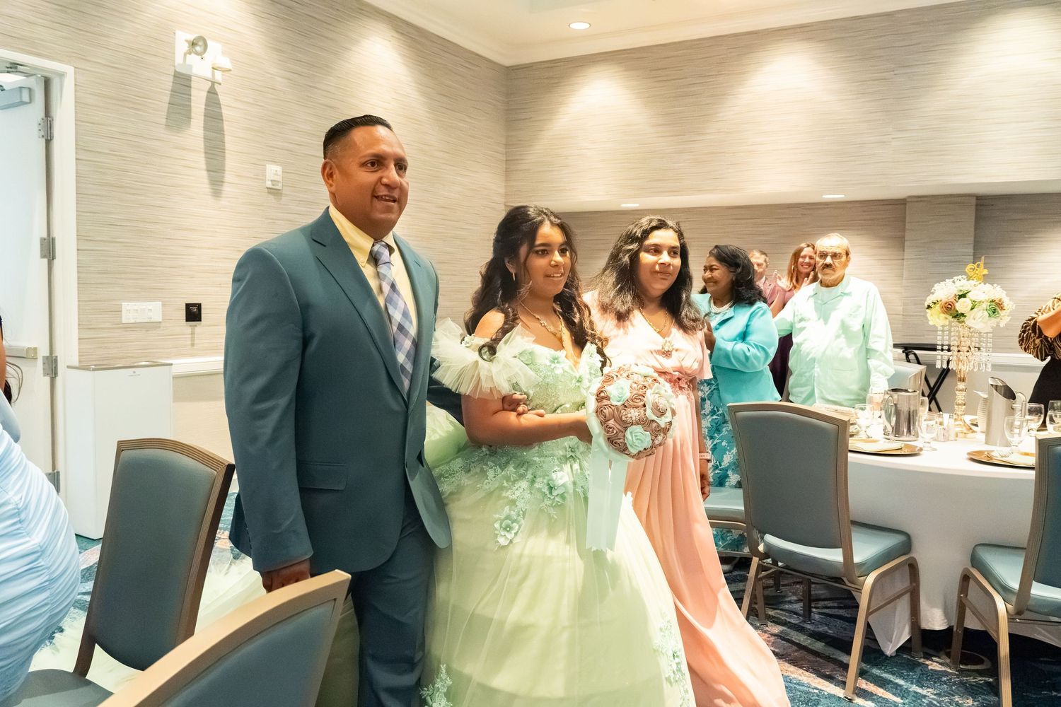 Man in blue suit escorts two women in mint green gowns at an elegant wedding reception venue.