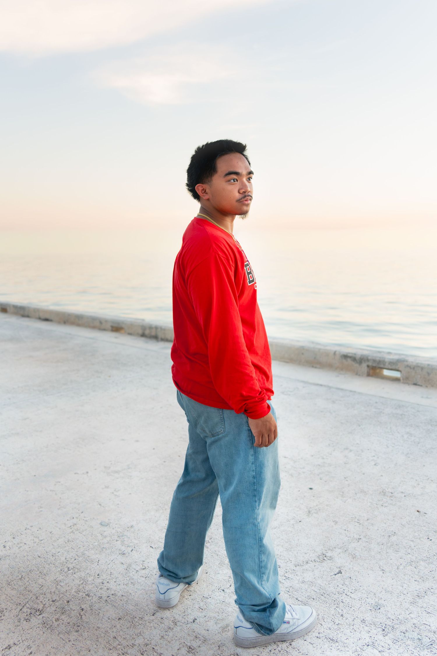 Young man in red sweatshirt and jeans walking along seaside promenade at sunset.