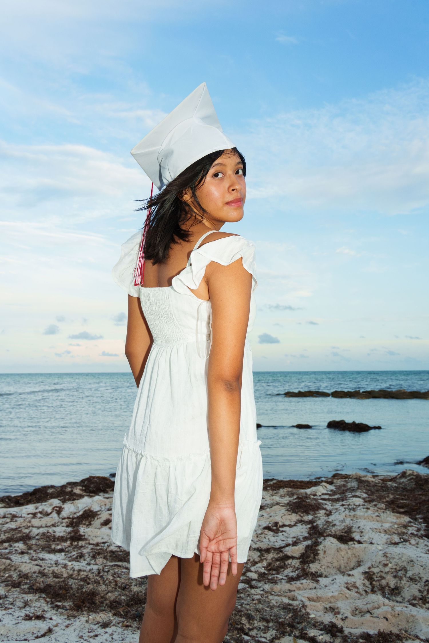 Young woman in white dress and graduation cap stands on rocky beach with ocean and blue sky behind her.