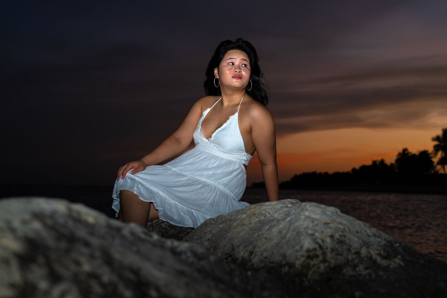 A figure in a white dress poses on rocks at sunset against a dramatic orange and purple sky.