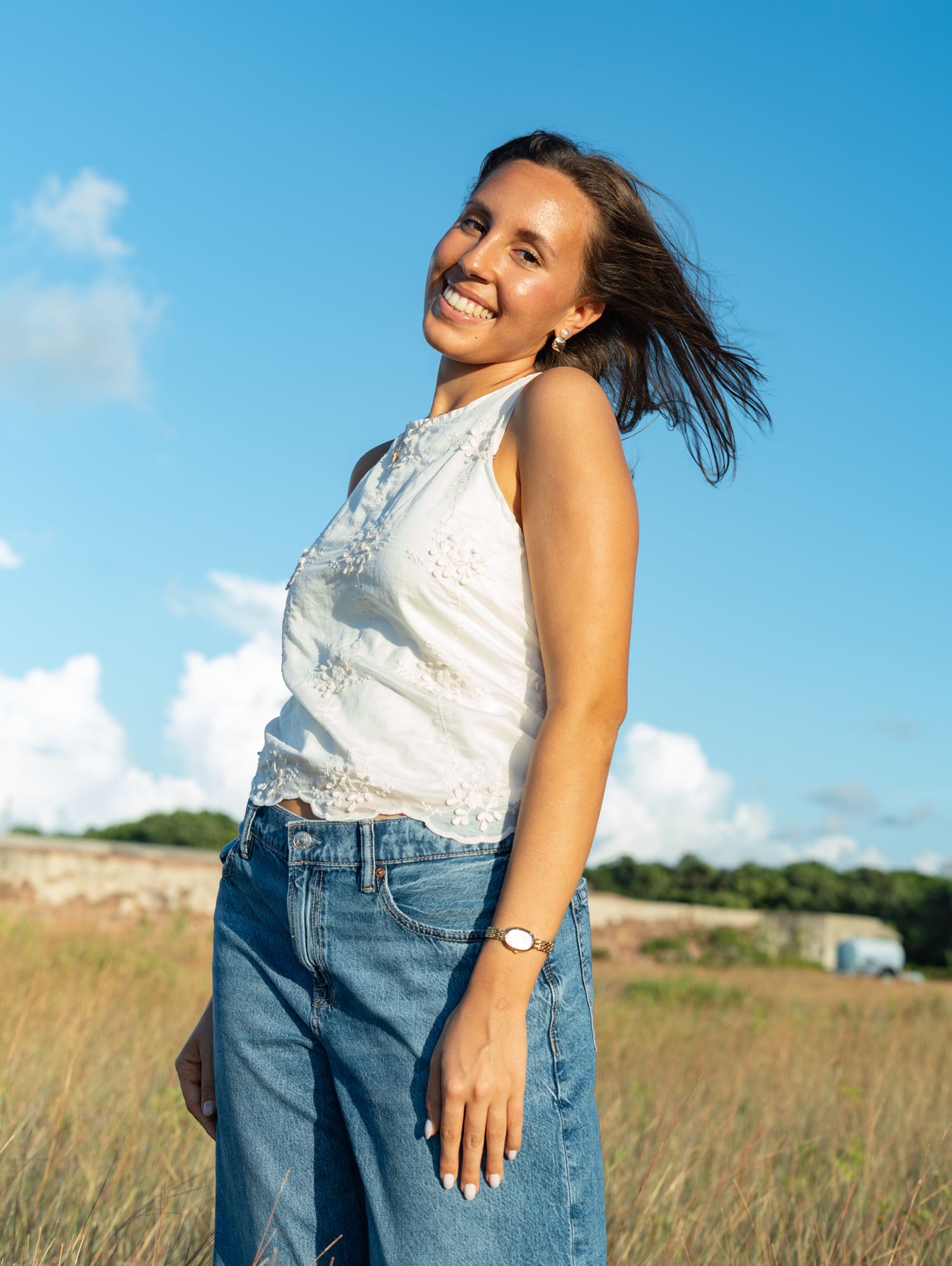 Someone in a white top and jeans stands joyfully in a grassy field against a bright blue summer sky.