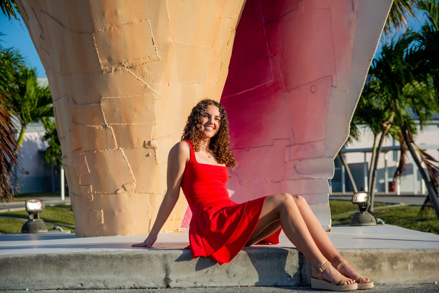 A person in a flowing red dress poses gracefully against a curved architectural structure in a tropical setting.