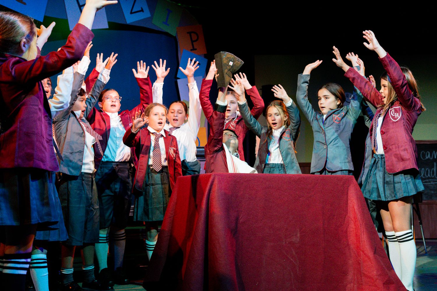 Students in school uniforms enthusiastically performing with raised hands during a stage production.