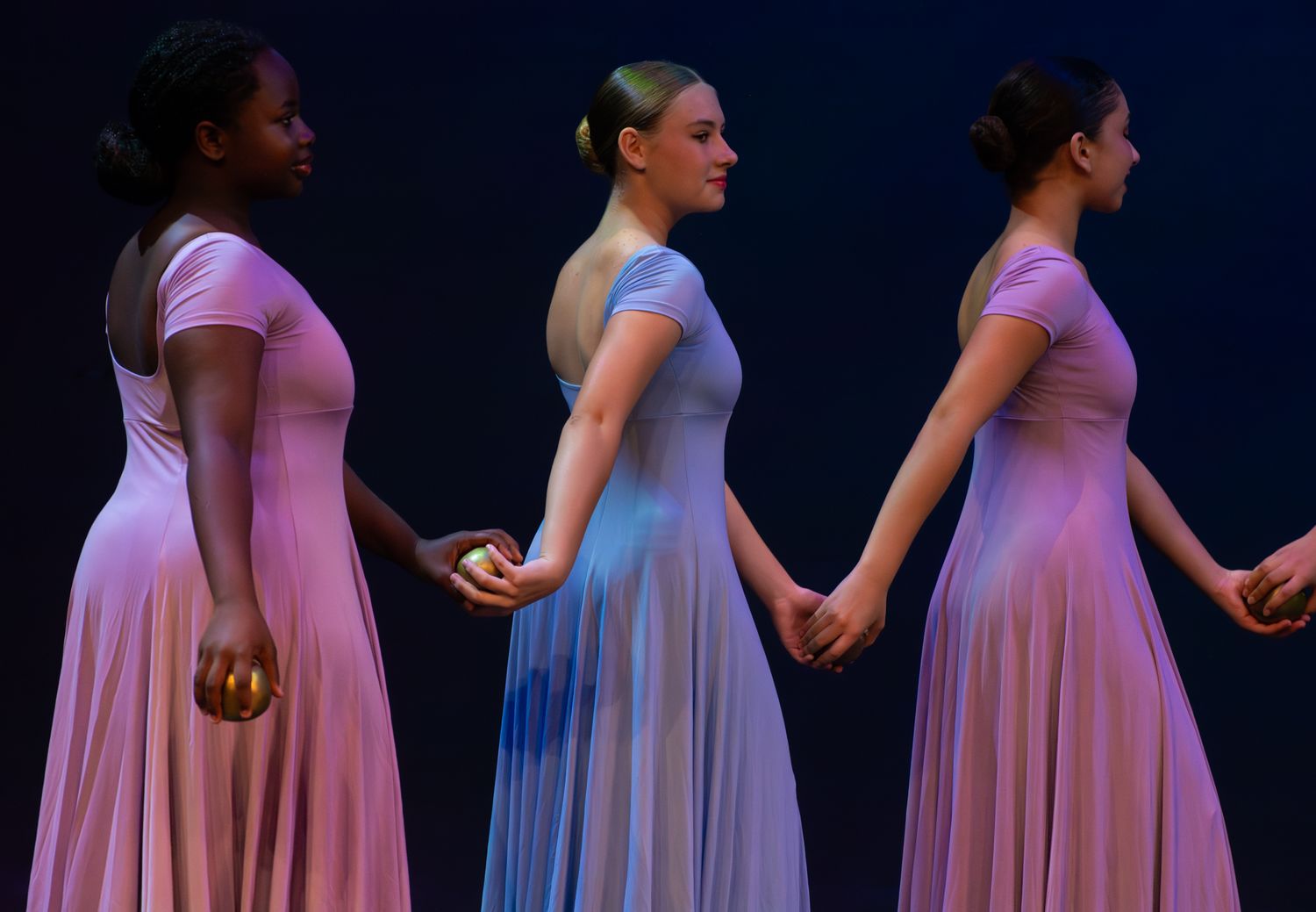 Three dancers in flowing pink and blue dresses perform synchronized movement on dark stage.
