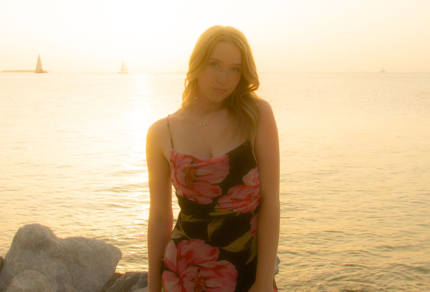 Woman in floral dress poses at sunset near ocean.