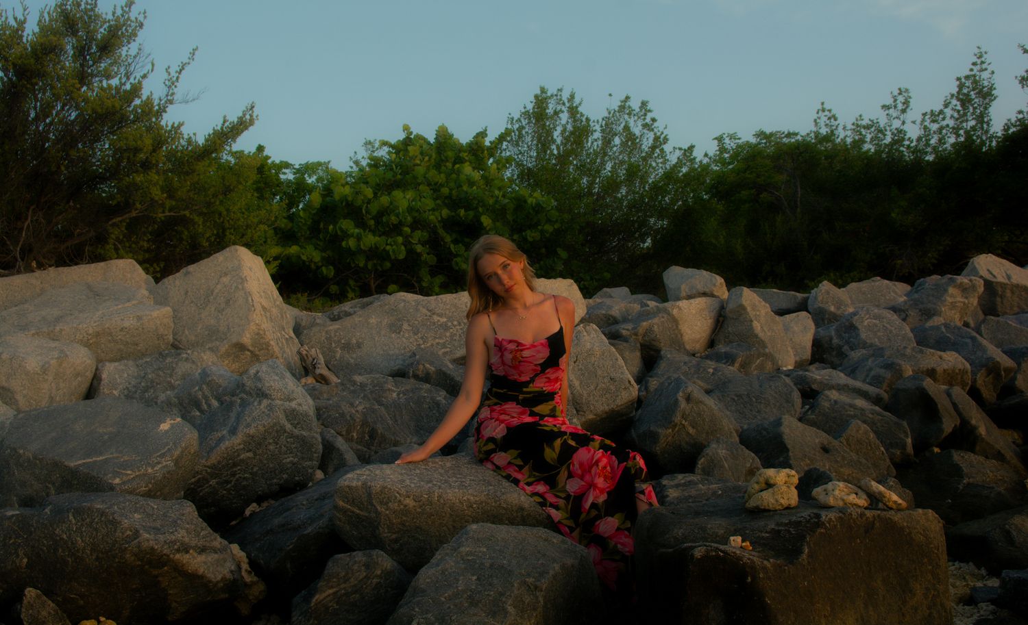 A figure in a floral dress poses on coastal rocks at sunset.