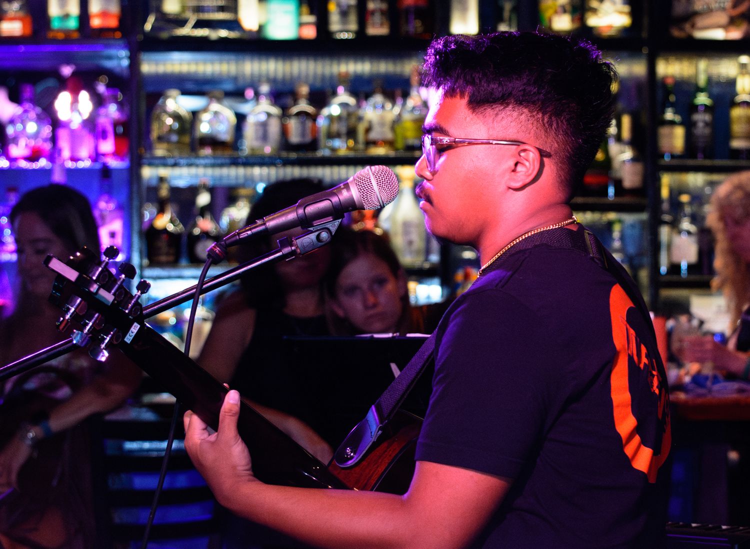 Musician performs with guitar and microphone in front of bar's neon-lit backdrop.