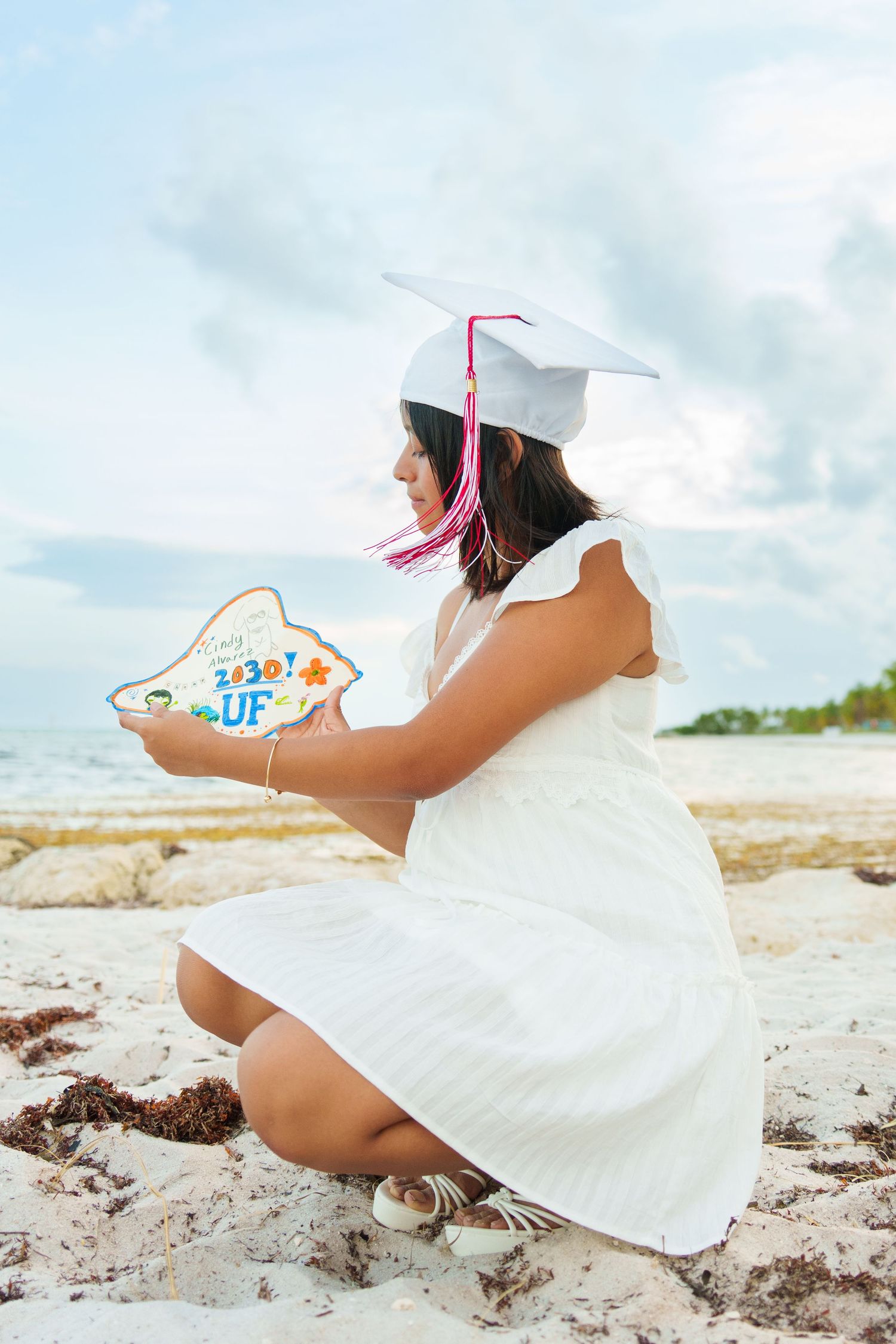 Graduate in white cap and dress crouches on sandy beach holding colorful diploma-shaped cookie.