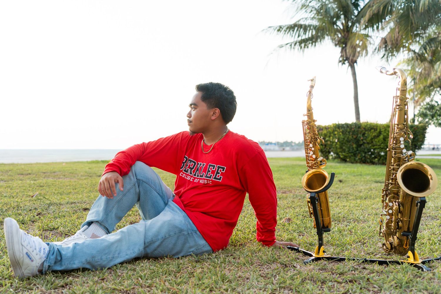 Young man in red sweatshirt sits on grass beside two saxophones near a tropical beach.