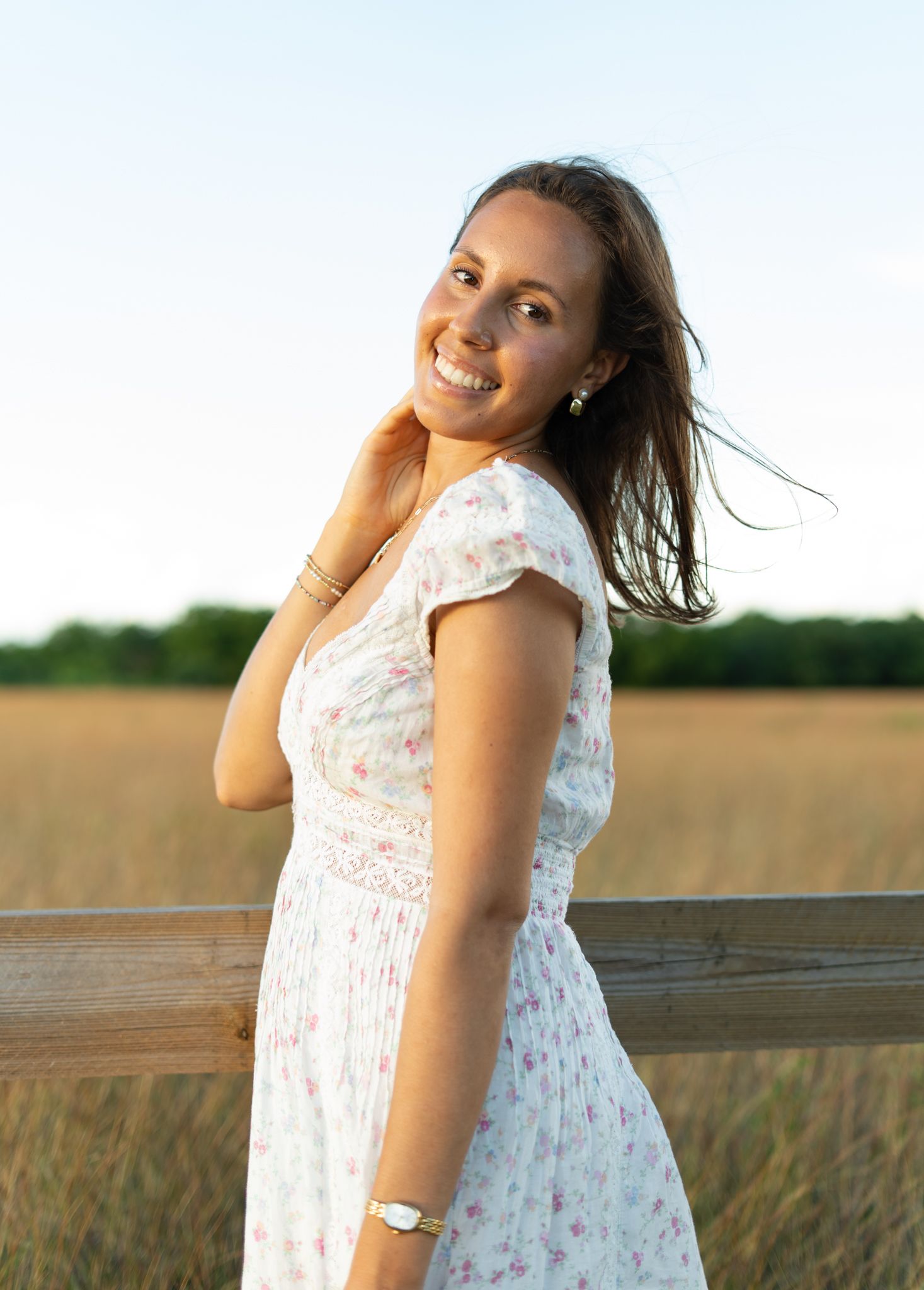 A woman in a white floral dress poses in a golden wheat field at sunset, creating a dreamy summer portrait.