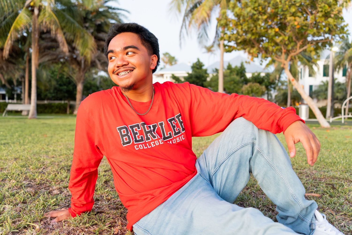 Young man in red Berklee College sweatshirt sitting on grass outdoors, smiling, with palm trees in background.