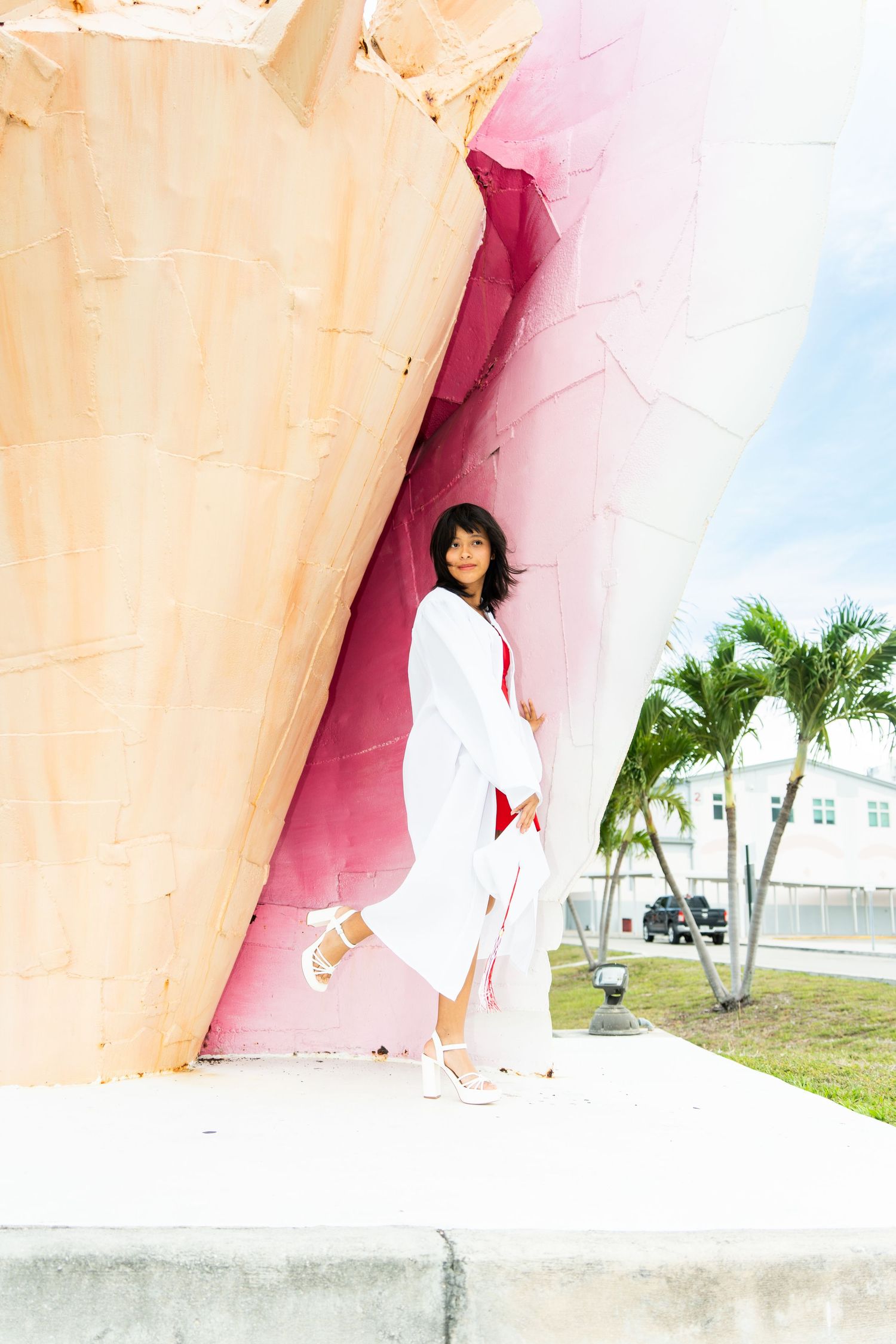 Graduate in white cap and gown poses beside a large pink decorative shell sculpture outdoors with palm trees.