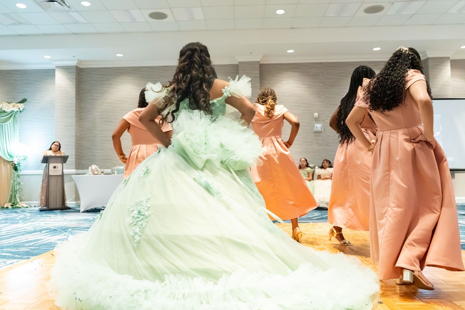 Young woman in mint green quinceañera gown twirls with bridesmaids in pink dresses during celebration.