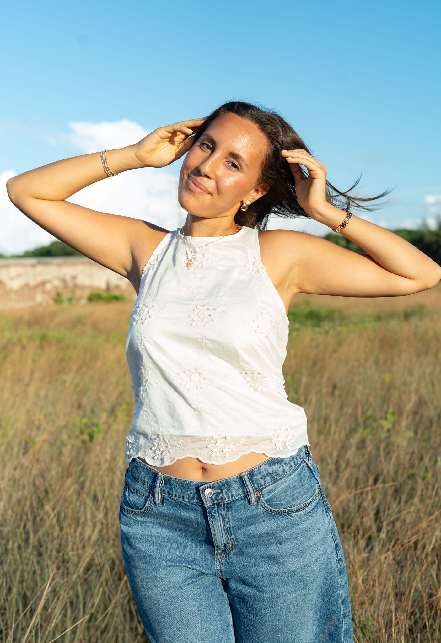 Person in white lace crop top and blue jeans poses playfully in a sunny field against bright blue sky.