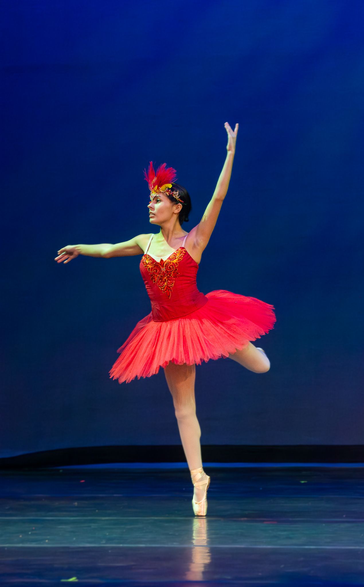 Ballerina in red tutu and feather headpiece performs arabesques pose on stage with blue backdrop.