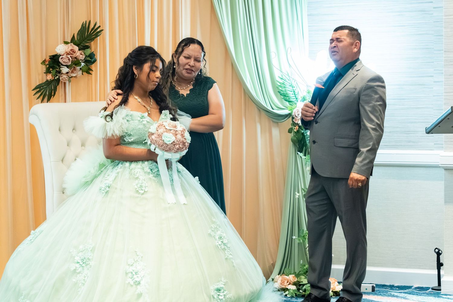 Young woman in mint green quinceañera gown holds bouquet beside two guests in elegant decorated venue.
