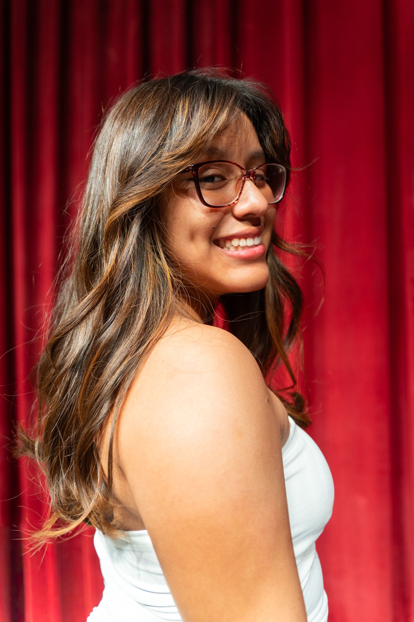Smiling young woman with long brown hair and glasses wearing a white dress against a red curtain backdrop.