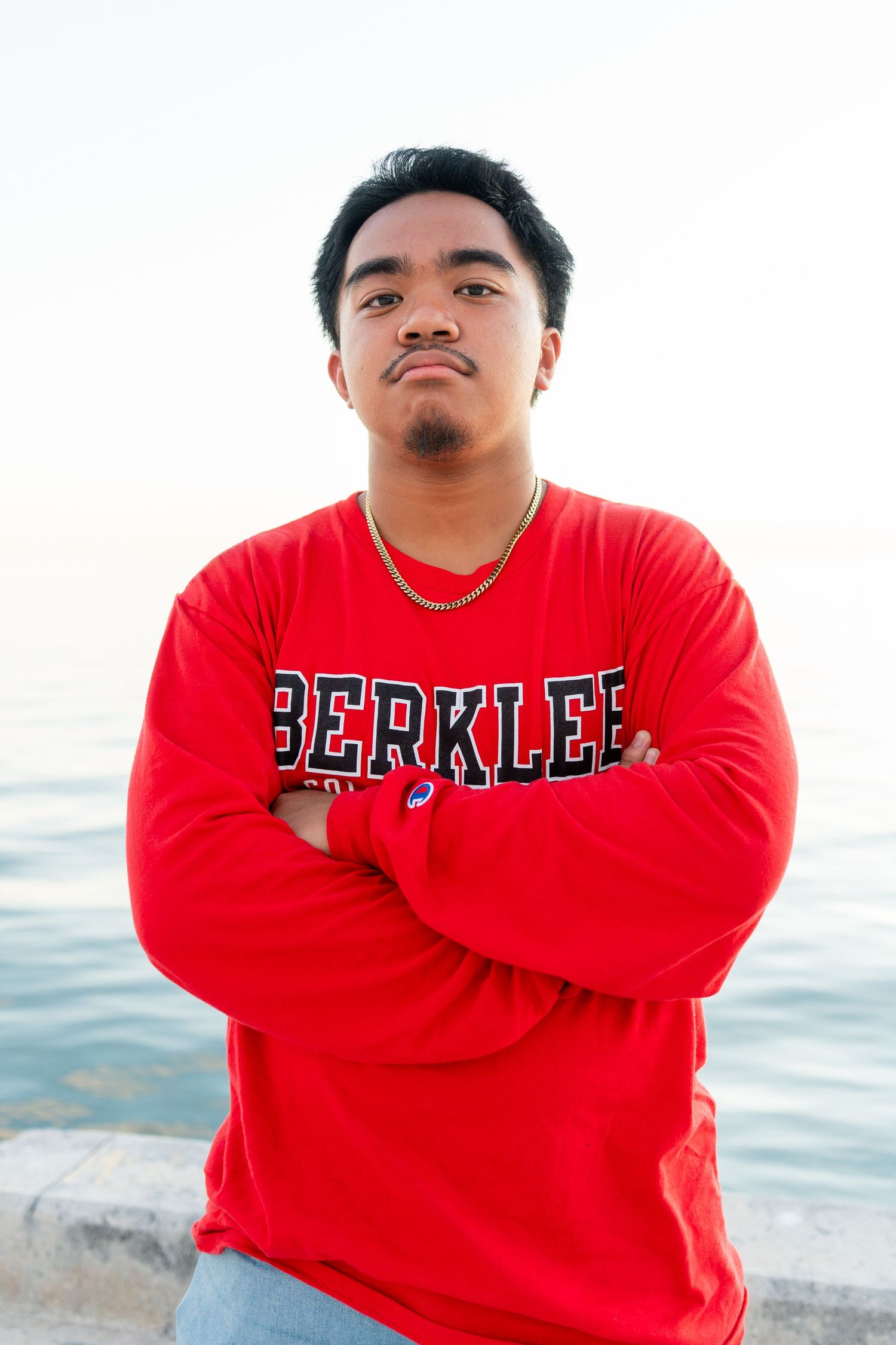 Young man in red Berklee sweatshirt stands arms crossed near waterfront, wearing gold chain necklace.