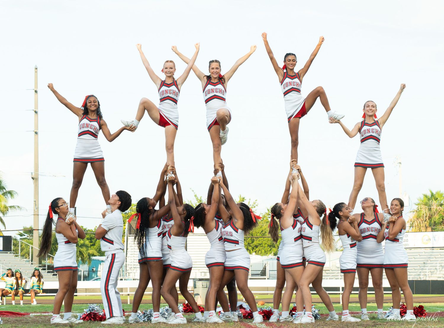 Cheerleaders in white and red uniforms perform a pyramid stunt on a field, with flyers raised high in formation.
