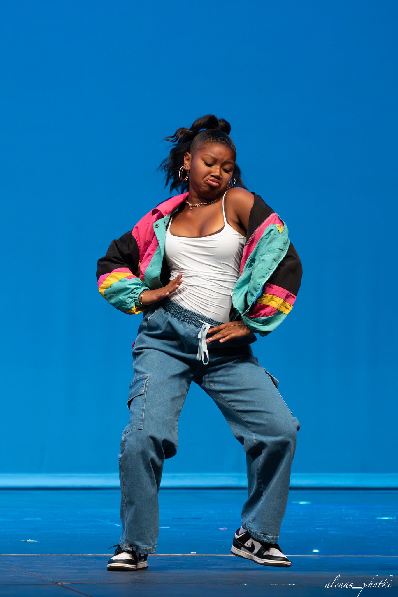 A young woman dances on stage in baggy jeans, white tank, and colorful jacket against a blue backdrop.