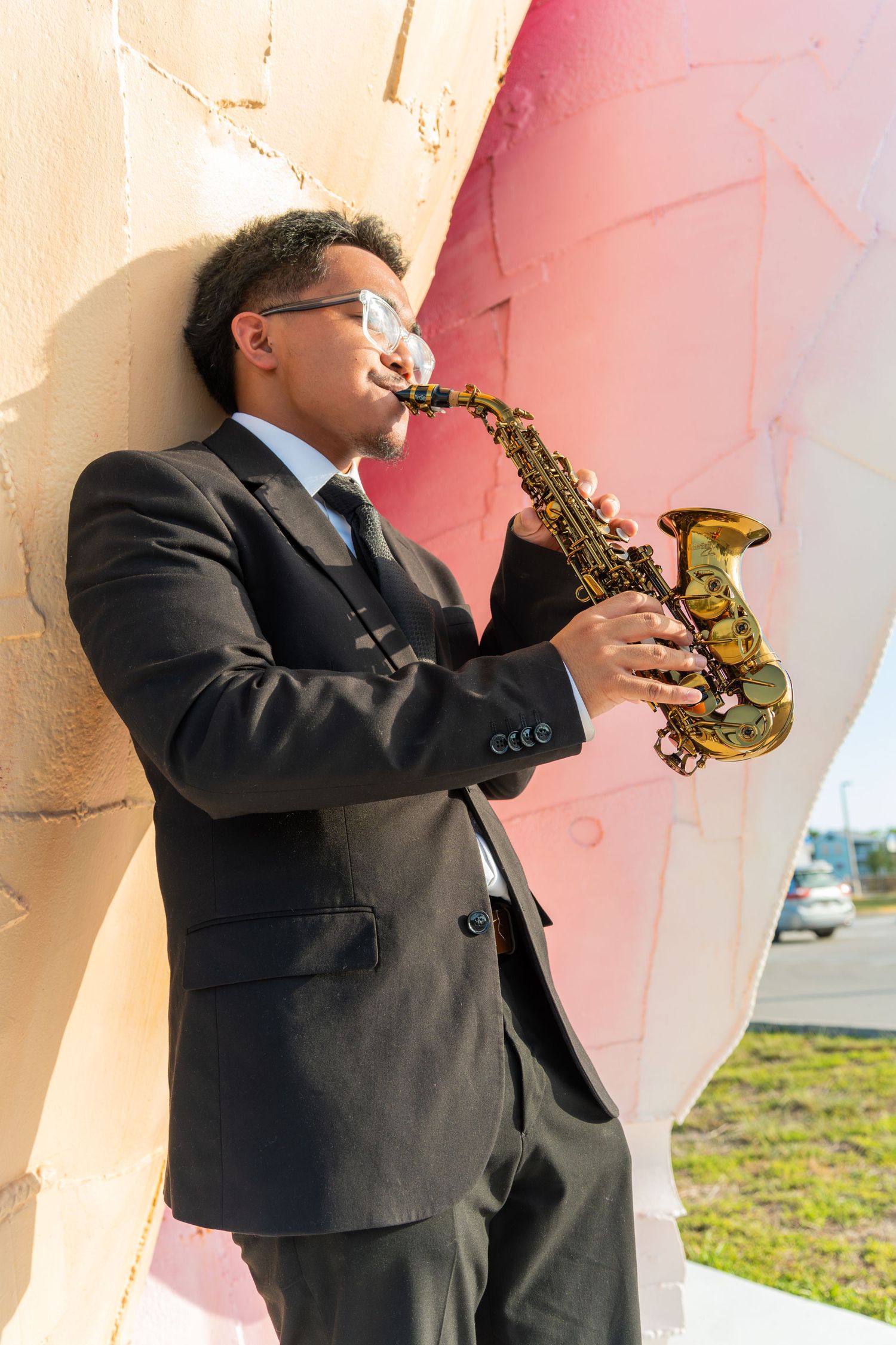 A man in a black suit plays a golden saxophone outdoors, leaning against a colorful pink and white wall.