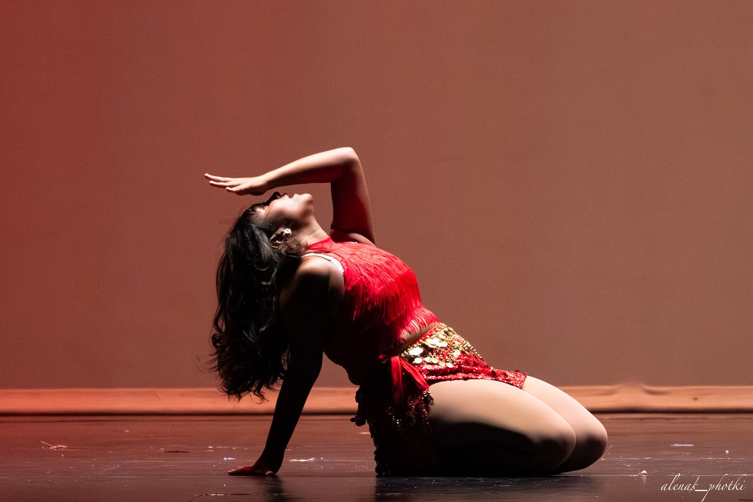 A dancer in a red costume performs an expressive floor pose on a dark stage under dramatic lighting.