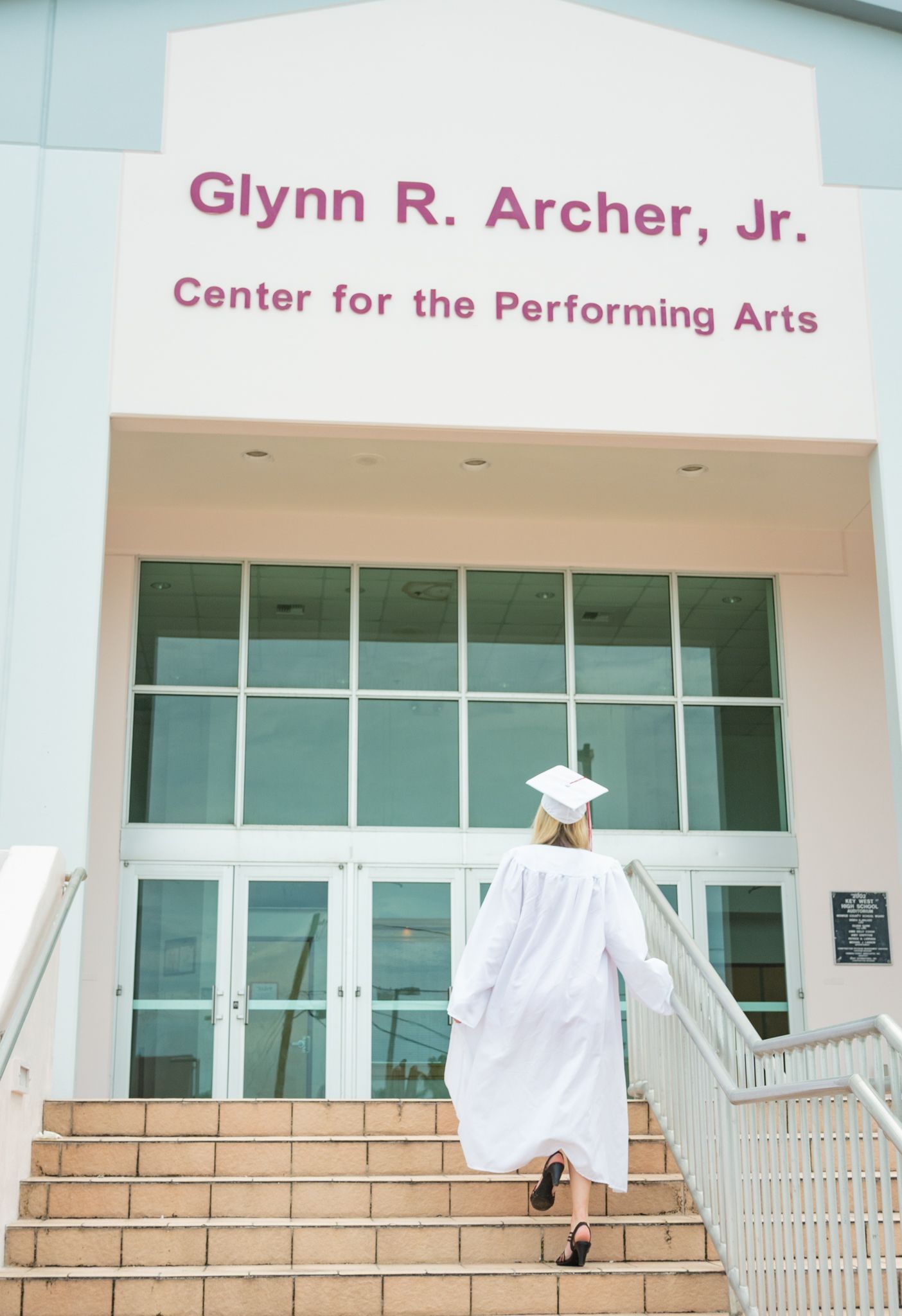 Graduate in white cap and gown ascending steps at Glynn R. Archer Jr. Center for the Performing Arts.