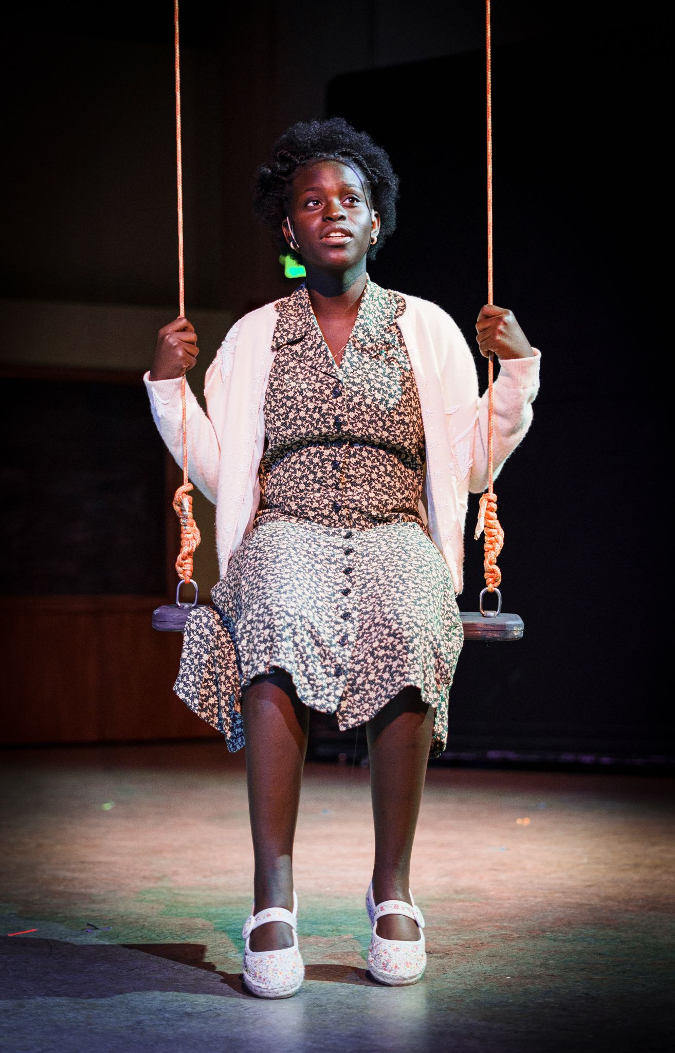Performer in vintage dress and cardigan sitting on a swing during a theatrical production.