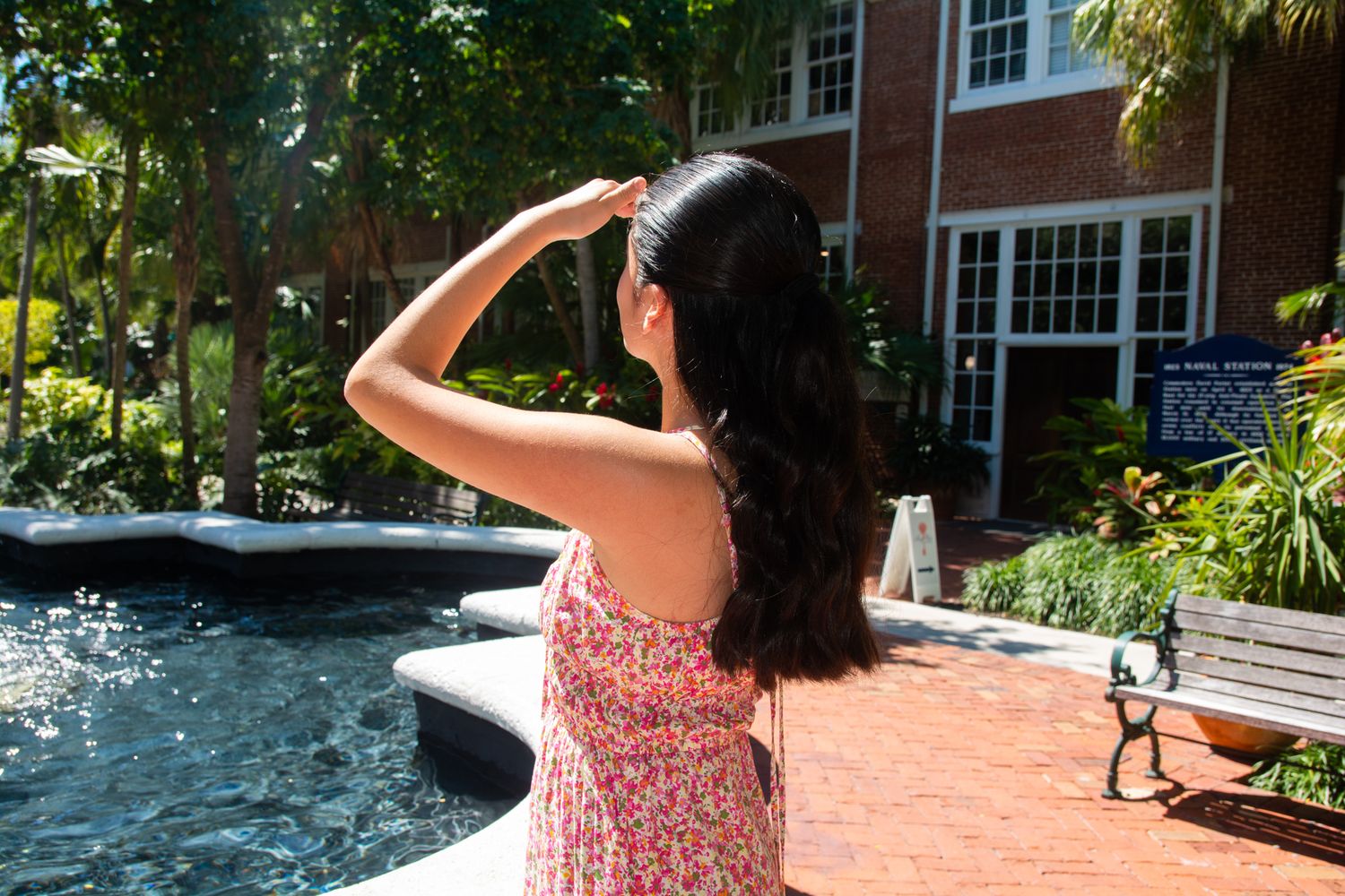 Graduate in a floral dress stands by a fountain near brick building on campus during sunny day.
