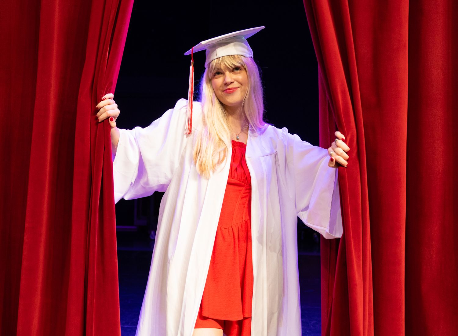 Woman in white graduation cap and gown over red dress, smiling between red curtains on stage.