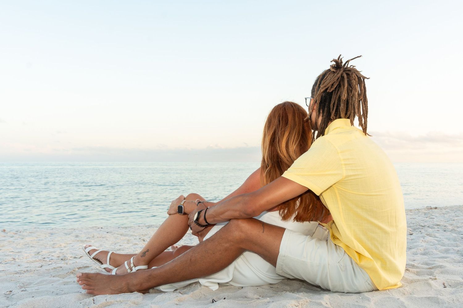 Couple sitting together on a sandy beach, embracing while gazing at the calm ocean during sunset.