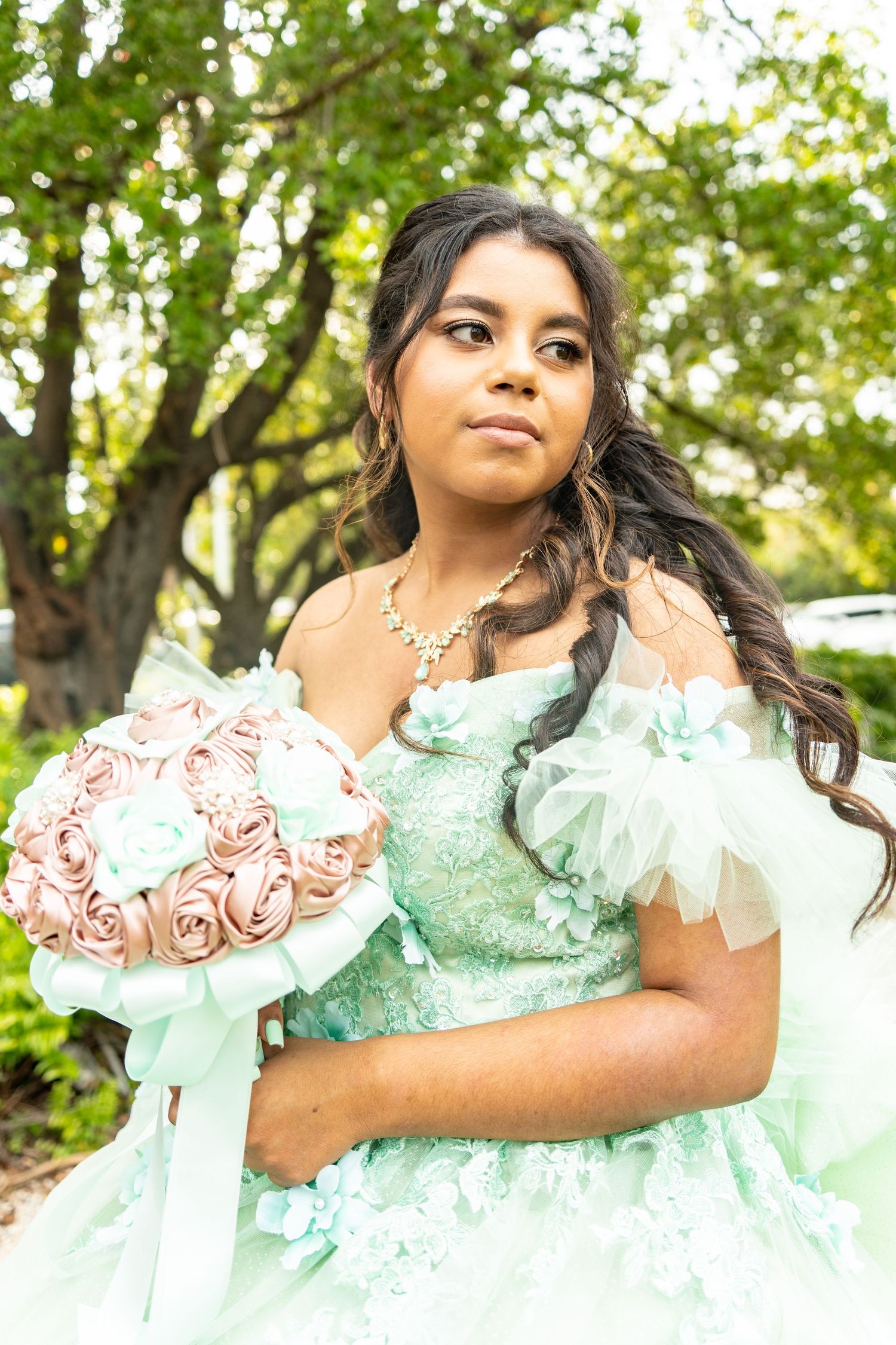 Young Latina girl in mint green quinceañera gown holds pink rose bouquet outdoors among lush green trees.