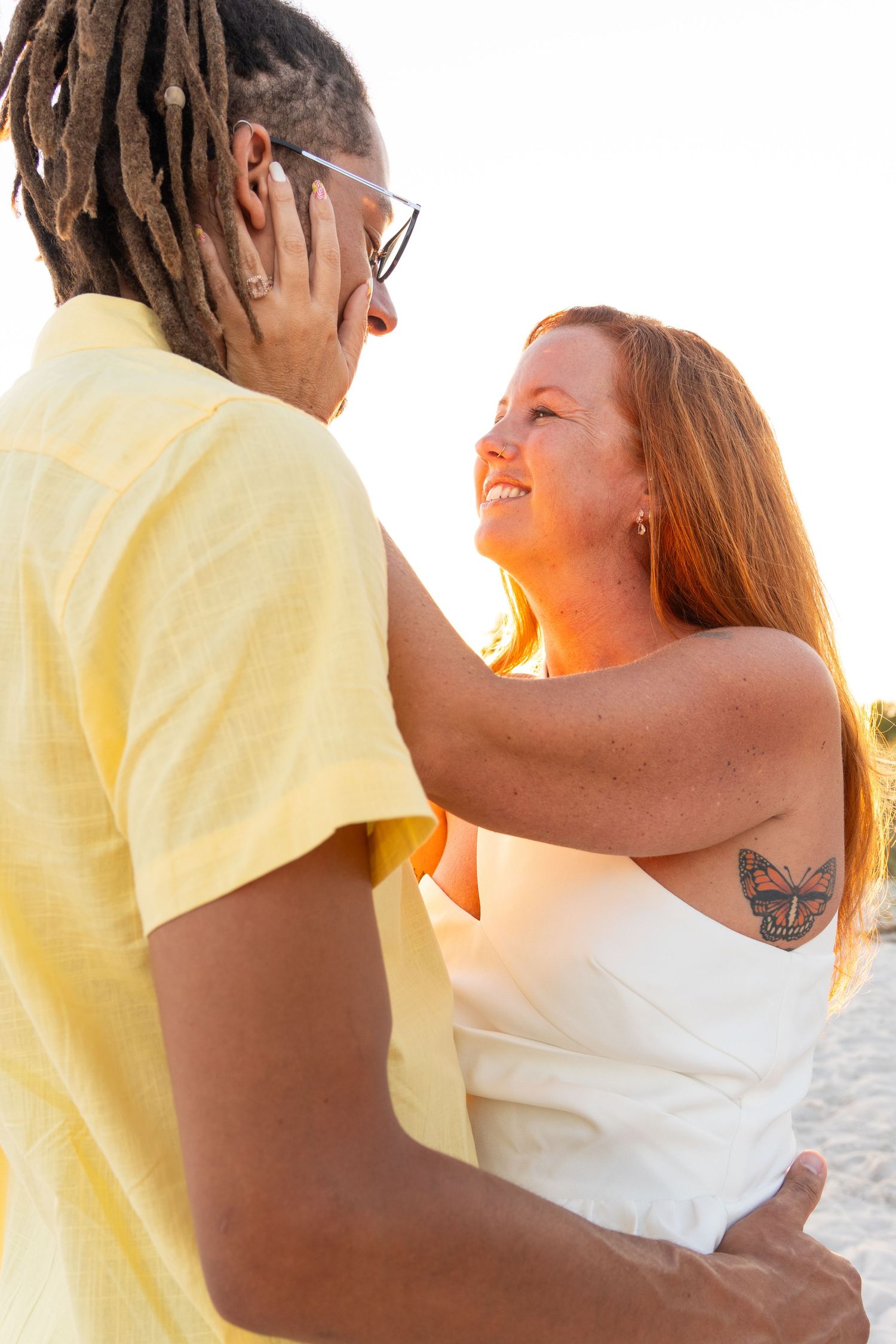 A smiling redhead woman with butterfly tattoo embraces a man with dreadlocks and glasses on a sunny beach.