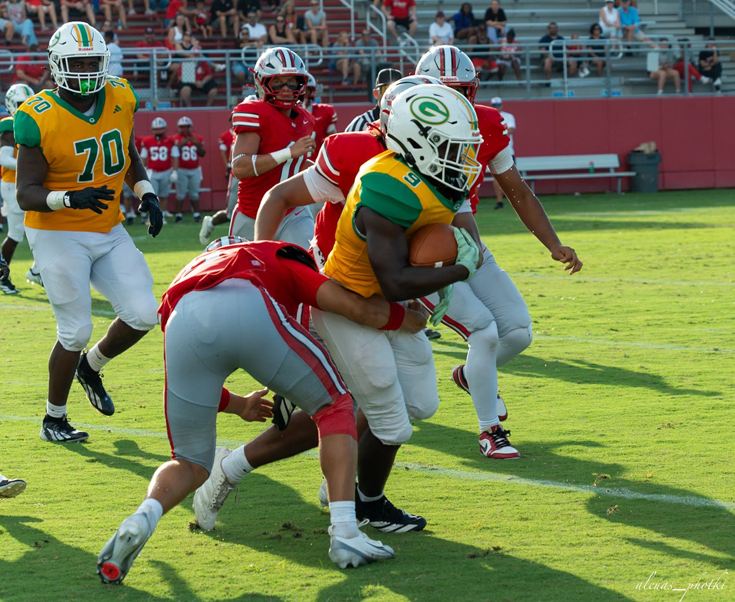 Football player in green and gold uniform runs with ball while being tackled by red team defenders.