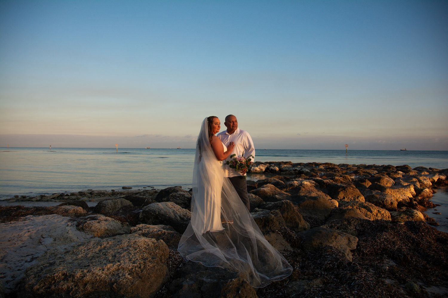 Newlyweds embrace on rocky shore during sunset with veil flowing in ocean breeze.