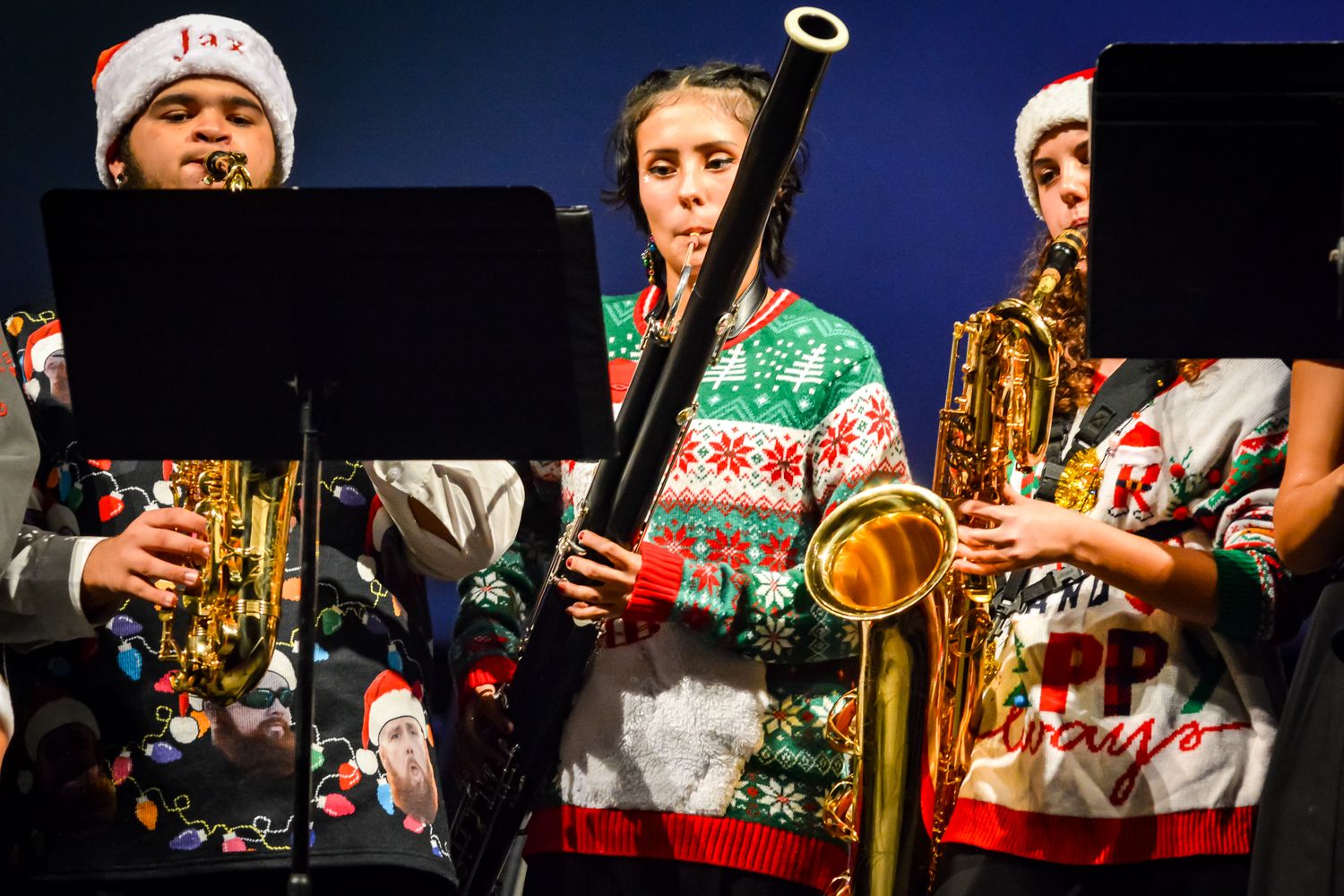 Student musicians wearing holiday sweaters perform with brass and woodwind instruments.