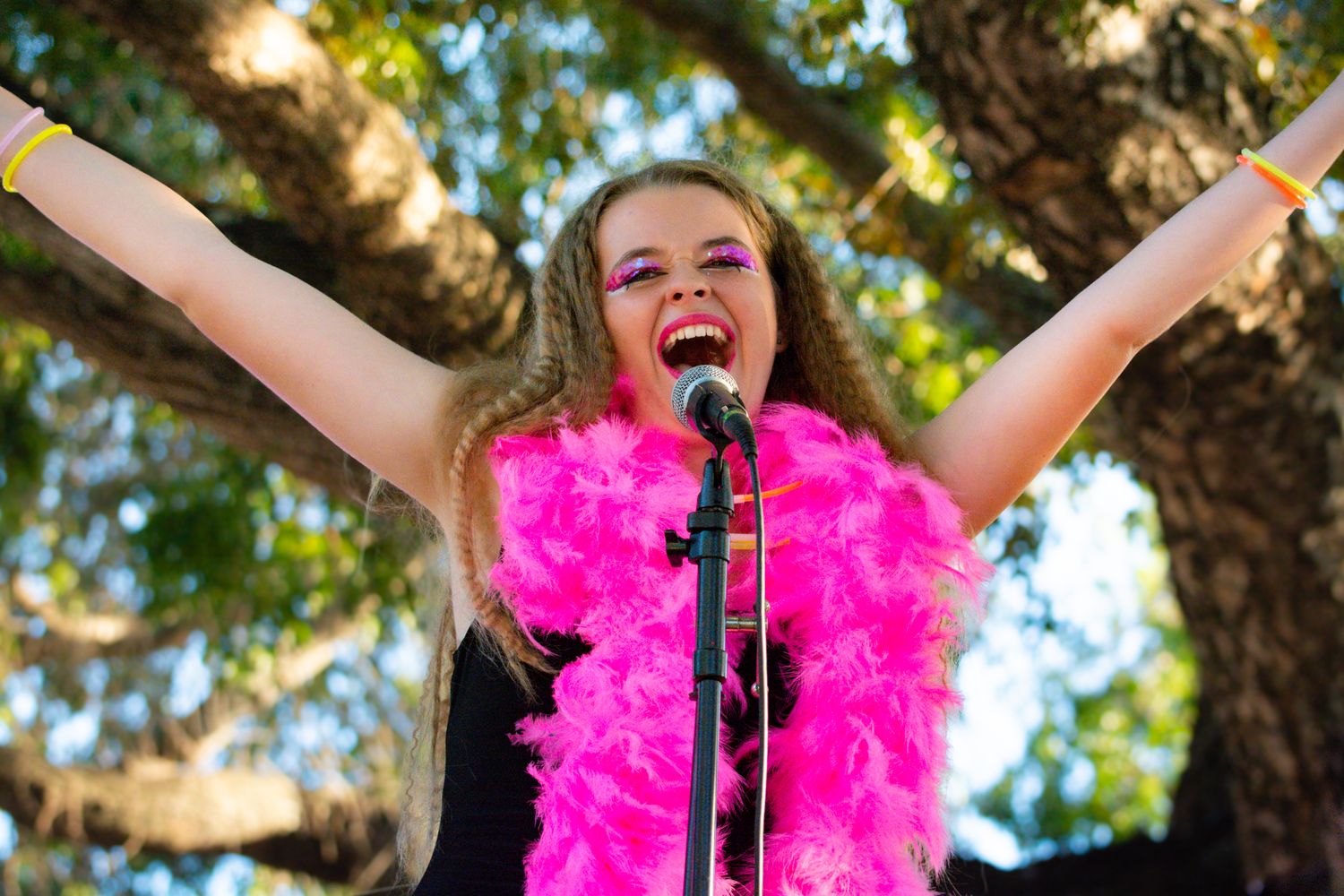 Singer performs enthusiastically with microphone and pink feather boa against outdoor tree backdrop.