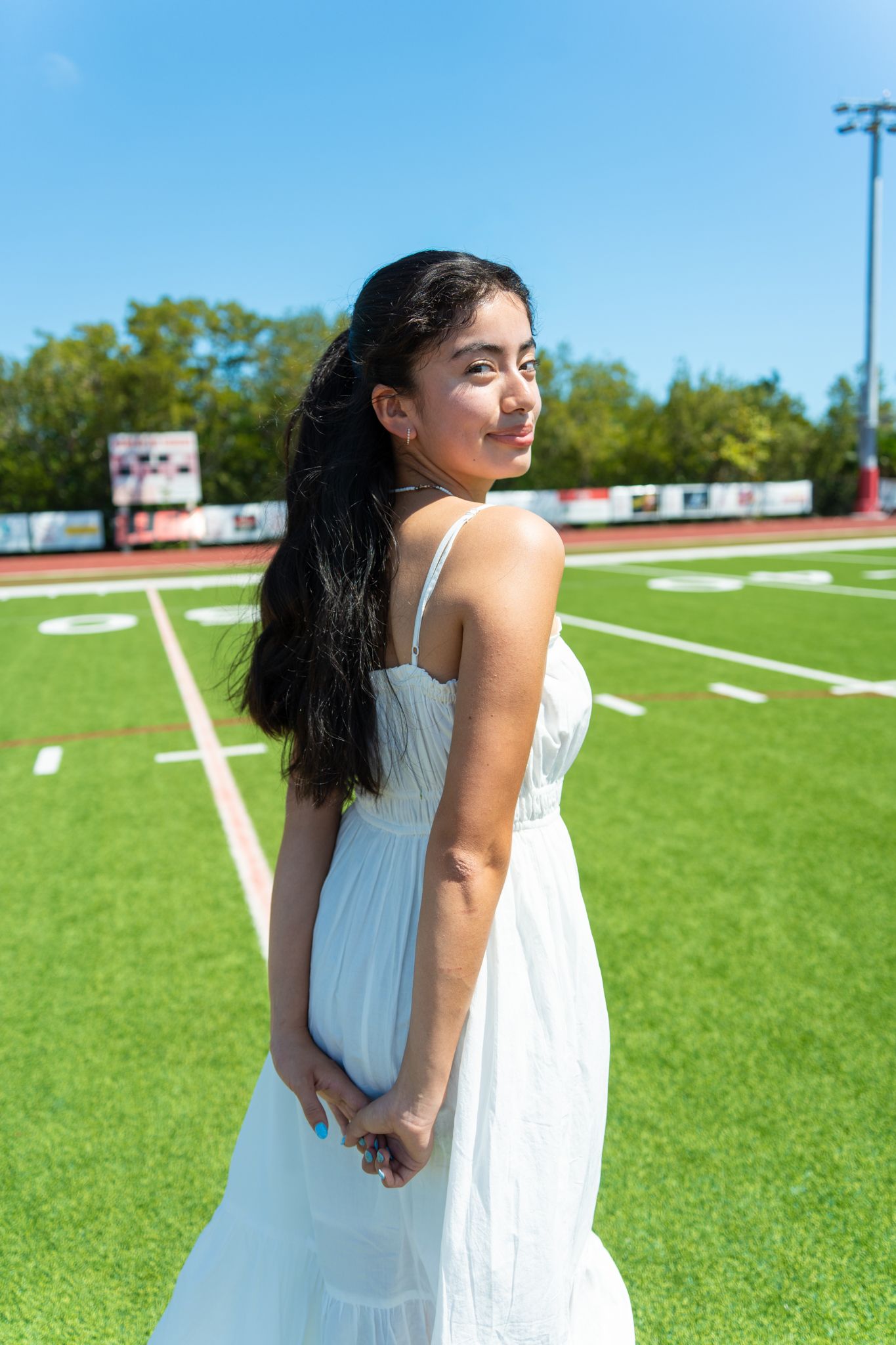 Young woman in white sundress poses on green football field under blue sky, looking back over her shoulder.
