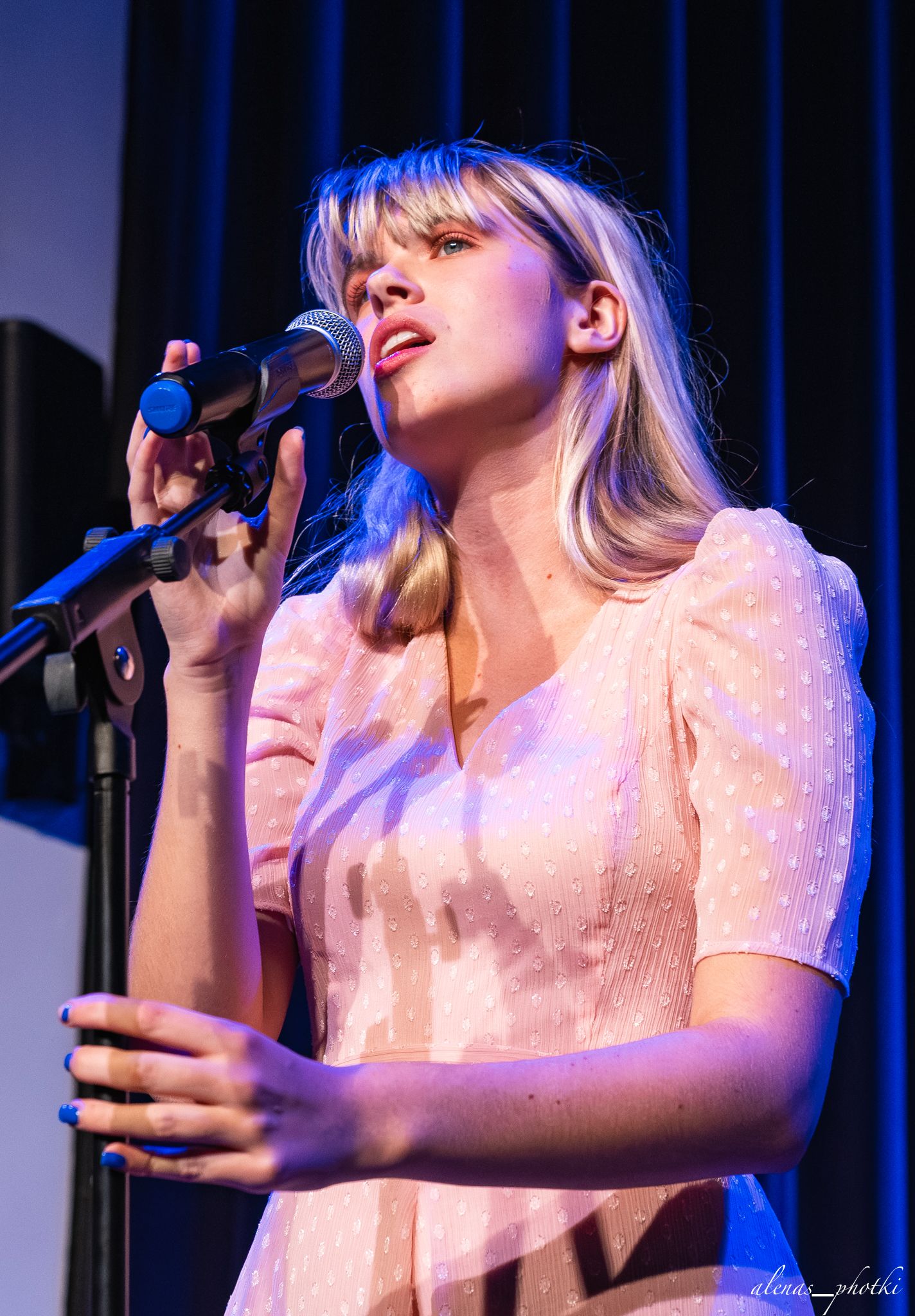 A performer in a white dress sings passionately on stage under blue lighting during a live musical performance.