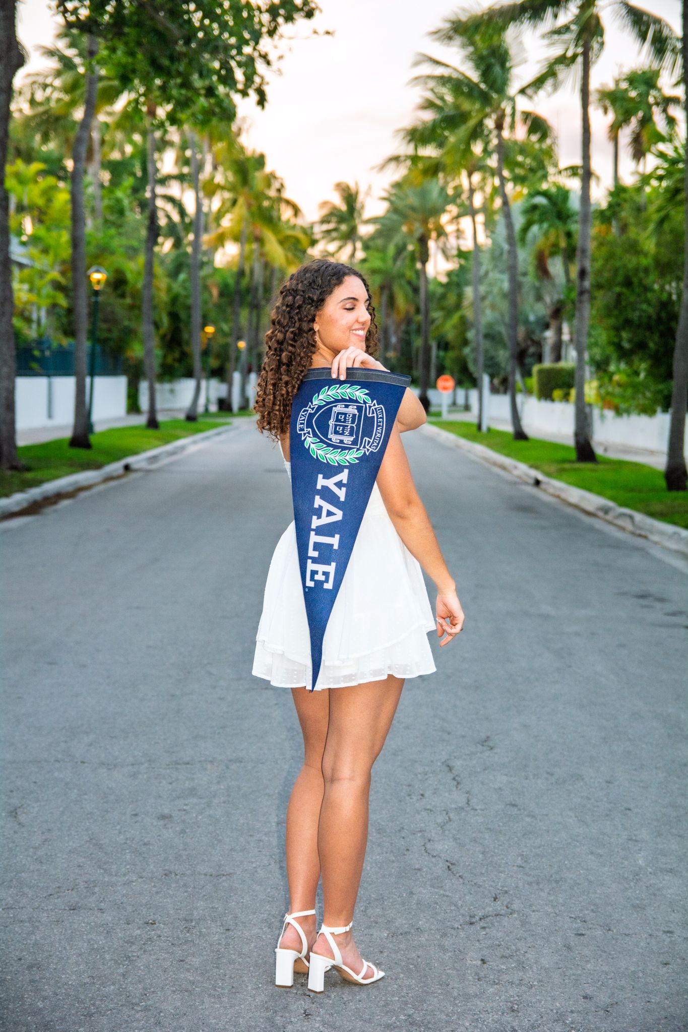 White dress and Yale pennant photographed on palm-lined street in elegant pose.