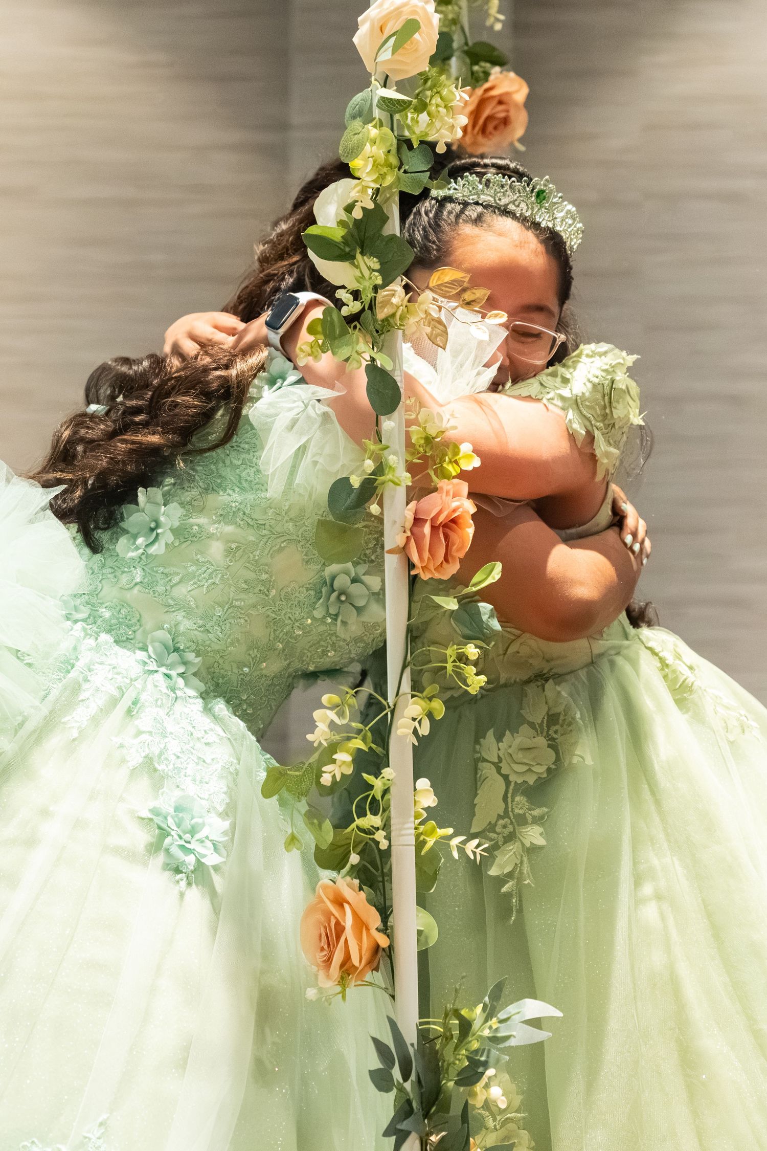 Two girls in mint green floral ball gowns pose with a flower-adorned pole at a quinceañera celebration.