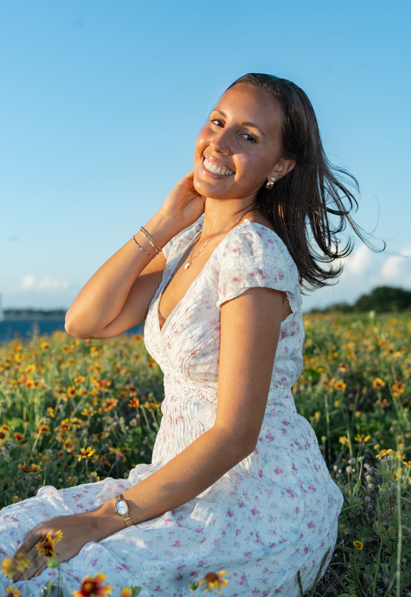 A white floral dress flows in the breeze as someone poses playfully in a sunflower field on a bright summer day.
