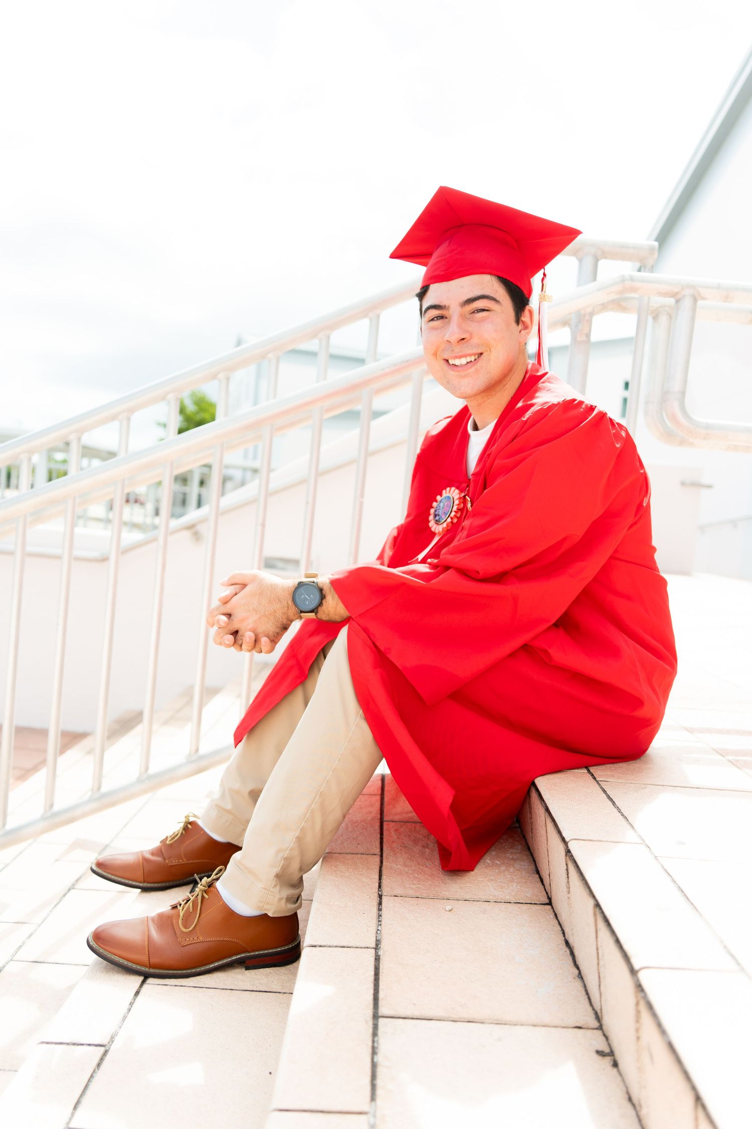 Smiling young man in red graduation cap and gown sitting on steps, wearing brown dress shoes and a watch.