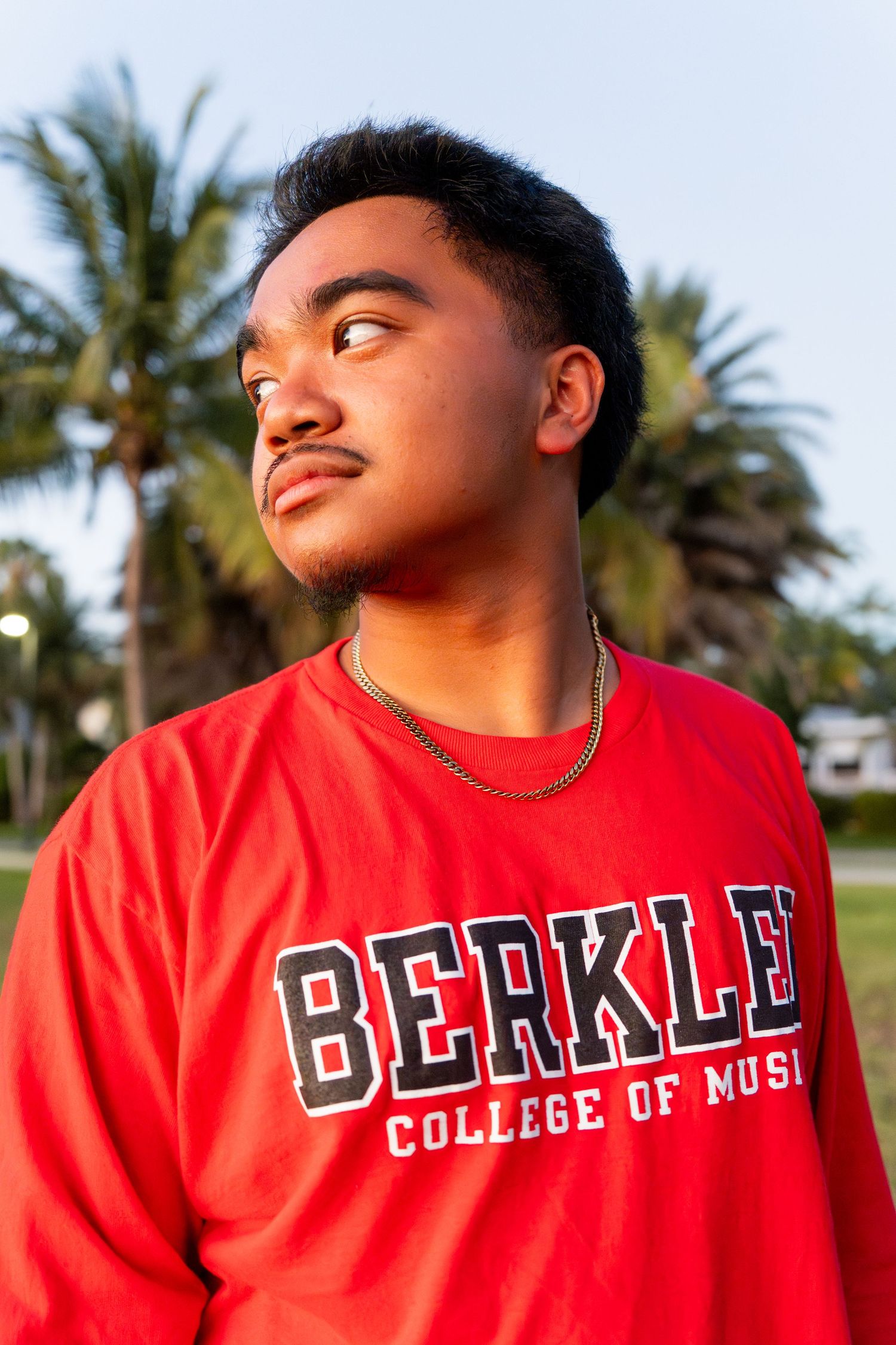 Young man wearing a red Berklee College of Music sweatshirt and gold chain, standing outdoors near palm trees.