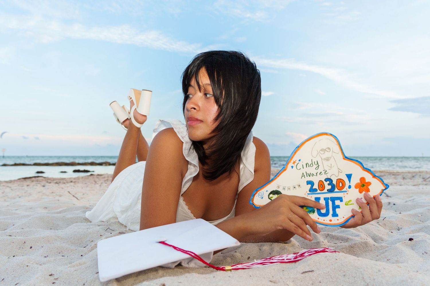 Young woman in white dress lying on beach sand holding a UF Class of 2020 graduation sign.