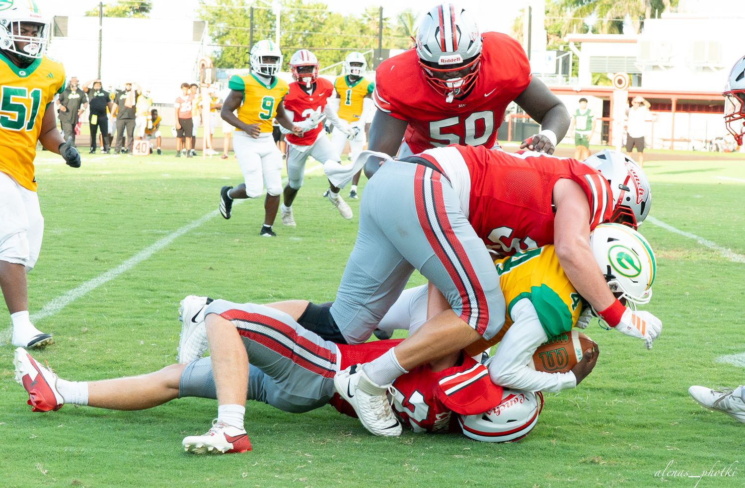 High school football players in red uniforms tackle opponent during competitive game on green field.